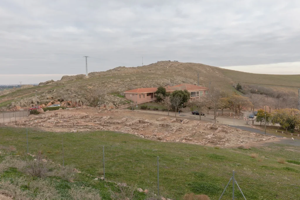 Restos excavados en el parque arqueológico de Alarcos, con edificios modernos y una loma al fondo.