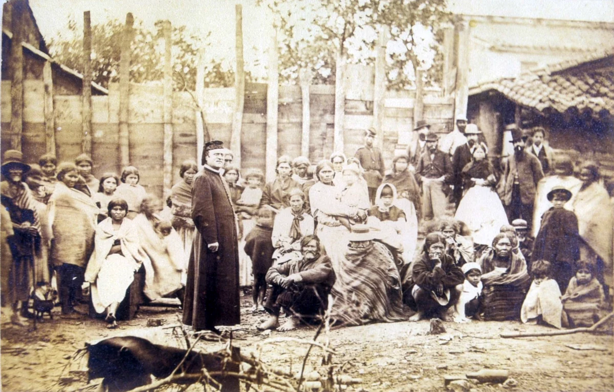 Grupo de adultos y niños refugiados de San Pedro, con un sacerdote en primer plano, durante la Guerra de la Triple Alianza.